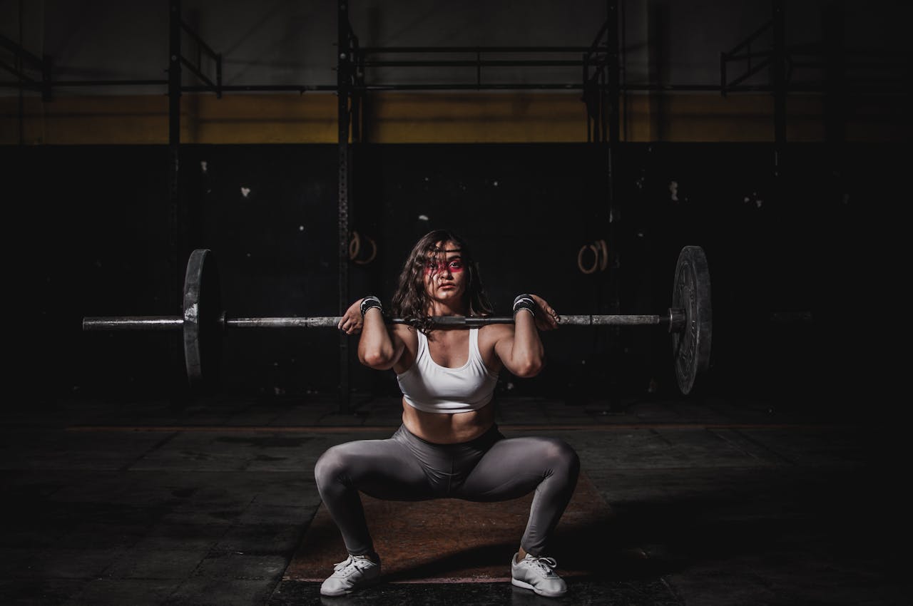 services-03 A strong female weightlifter squats with a barbell in a dimly lit gym, showcasing power and fitness.